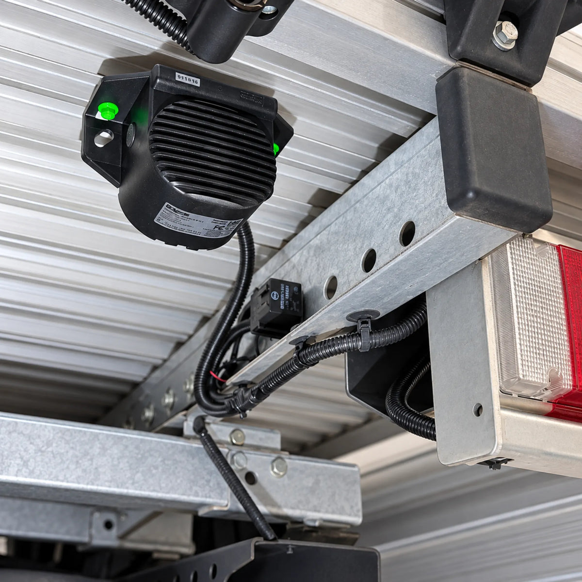 Brigade SA-BBS-97 reverse alarm installed upside down under the rear of a work vehicle tray, bolted to a galvanised steel crossmember beside the tail light, with the lead run in black corrugated conduit