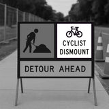 Cyclist Dismount sign panel mounted on a temporary sign frame next to a Works Ahead panel with Detour Ahead text, and traffic cones in the background