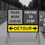 Temporary traffic control road work ahead sign with detour instructions on a stand.