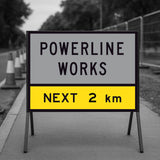 Temporary Traffic Control Warning sign indicating 'Powerline Works Next 2 km' on a road with traffic cones.