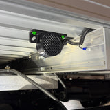 Brigade BBS-82 reverse alarm fitted under the rear of an aluminium truck tray, black mesh grille facing rearwards, with the wiring run in corrugated conduit along the chassis rail