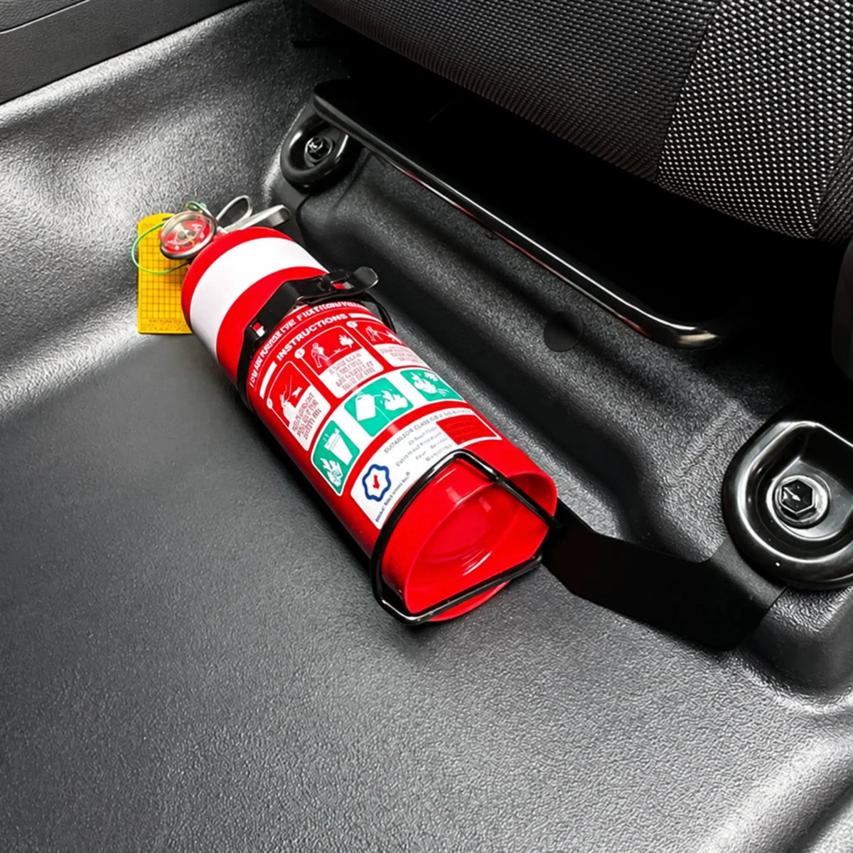 Red fire extinguisher held in its own wire strap bracket, bolted to a black seat mount bracket on the passenger seat rail of an N80 Toyota Hilux