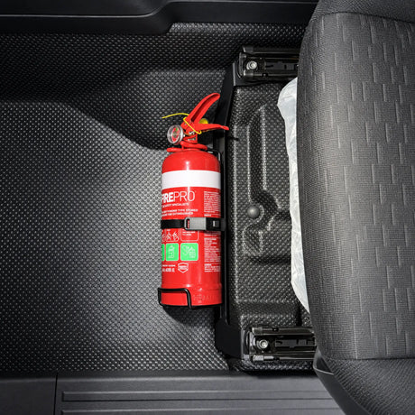 Red 1kg fire extinguisher held in its own strap bracket, bolted to a black seat mount bracket on the passenger seat rail of an MV Mitsubishi Triton, viewed from above