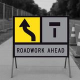 Lane Shifts Left TM10-15A corflute panel in a multi-message sign frame with the shift arrow in the left-hand position and a Lane Closed Ahead T symbol in the right-hand position, reading as the left lane shifting left with the right lane closed ahead, and a ROADWORK AHEAD panel below, on a footpath with traffic cones and temporary fencing