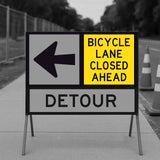 Bicycle Lane Closed Ahead sign panel mounted on a temporary sign frame with a Detour arrow panel and traffic cones in the background